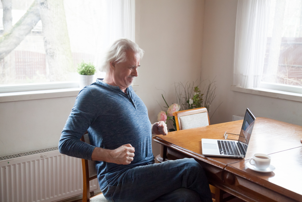 Grey haired middle aged senior male sitting alone in the kitchen on chair stretching back after long time working on computer doing exercises for loins preventing osteoarthritis and spinal stenosi