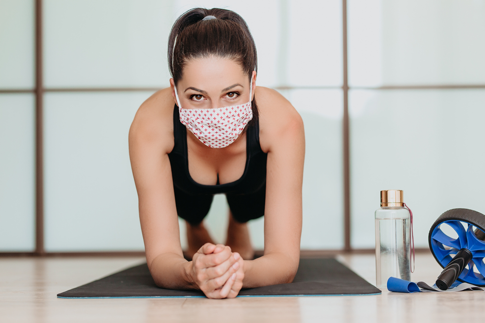 Getting fit during coronavirus pandemic - pretty fitness woman exercising at home during home quarantine, wearing medical face mask